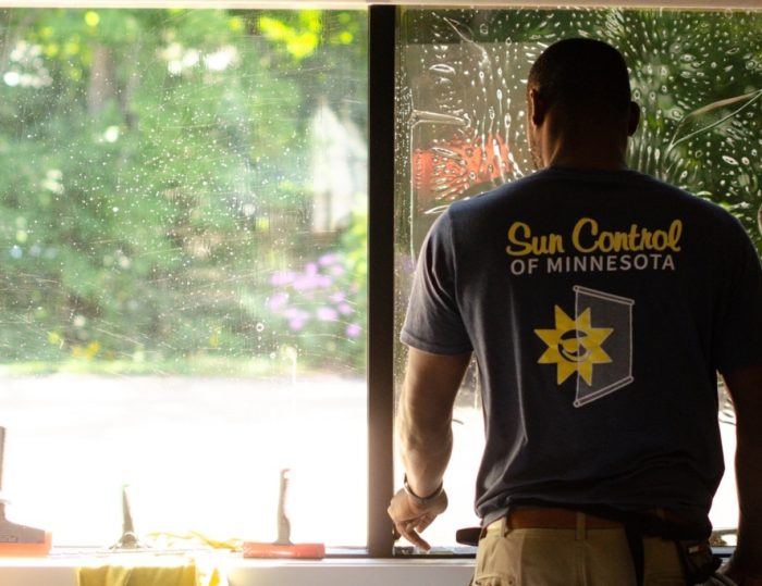 A technician from Sun Control of Minnesota stands at a residential window mid-installation, with one pane covered in soapy solution and tint film. Tools are lined up on the windowsill, illustrating the detailed and professional process of residential window tinting in a Minnesota home.